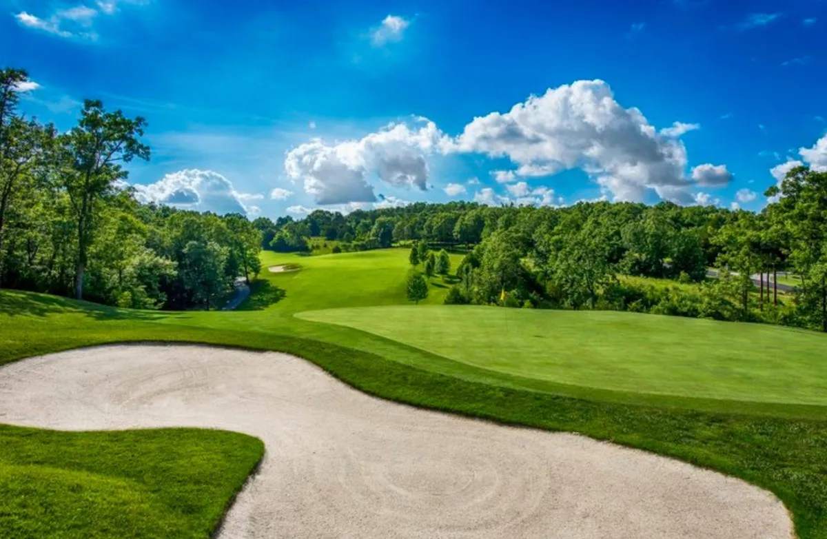 A sweeping exterior view of a lush, green golf course under a blue sky, with a sand trap in the foreground, mature trees lining the fairway, and gentle clouds above. The setting is vibrant and clearly designed for recreational golfing activities.