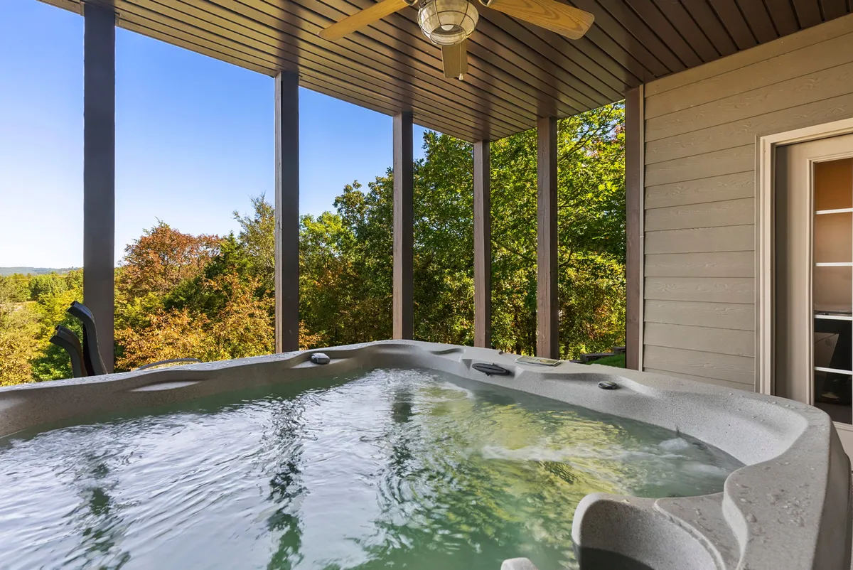 The image showcases the inviting outdoor hot tub set on a spacious deck, surrounded by lush trees that create a sense of privacy and tranquility. The ceiling fan adds a touch of comfort, and the setting sun bathes the area in a warm glow, hinting at the perfect spot for sunset views and stargazing. The overall mood is peaceful and welcoming, highlighting the natural beauty of the gated resort community.