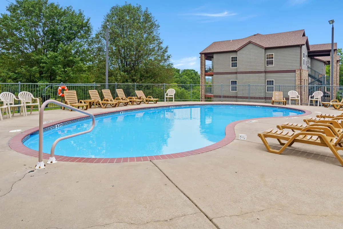 A swimming pool area with sparkling blue water, poolside chairs, and a backdrop of trees and a nearby building. The environment is relaxed, suggesting both active pool days and peaceful sunbathing—inviting for all ages.