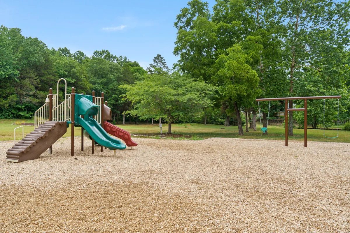 A lively playground scene surrounded by mature trees, with children swinging, playing, and families gathered nearby. The setting is joyful and energetic, capturing the spirit of family-friendly fun within the StoneBridge Resort.