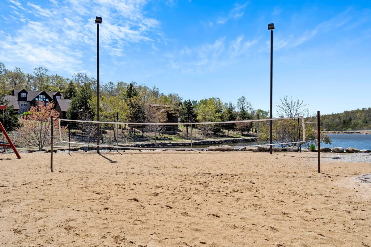 A lively exterior scene featuring a sandy volleyball court set along a waterfront, fringed by trees. People are seen engaged in a friendly game of volleyball, with the tranquil lake and wooded shoreline providing a picturesque, inviting backdrop for summer activities and relaxation.