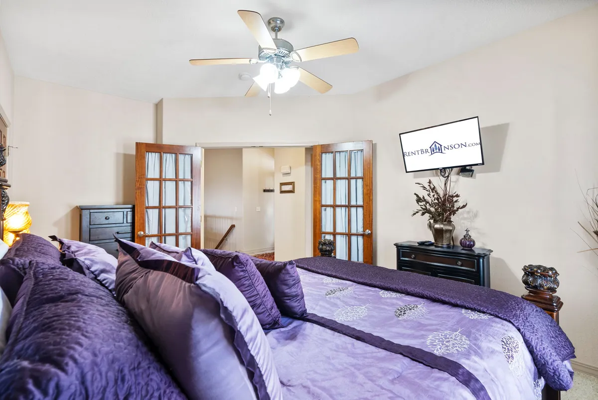 This inviting bedroom features plush purple bedding, elegant wooden furniture, a TV mounted on the wall, and a decorative plant. Natural light softly fills the room, creating a cozy yet sophisticated atmosphere ideal for restful nights.