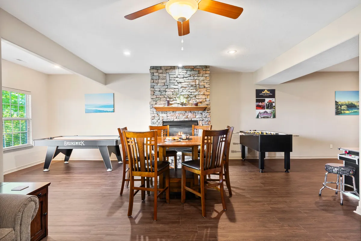 The image features a warm, playful interior scene centered on a dining table, with a fireplace and both air hockey and foosball tables nearby. The setup creates a lively environment suited for game nights, casual dining, and social gatherings, with a cozy atmosphere enhanced by the fireplace.