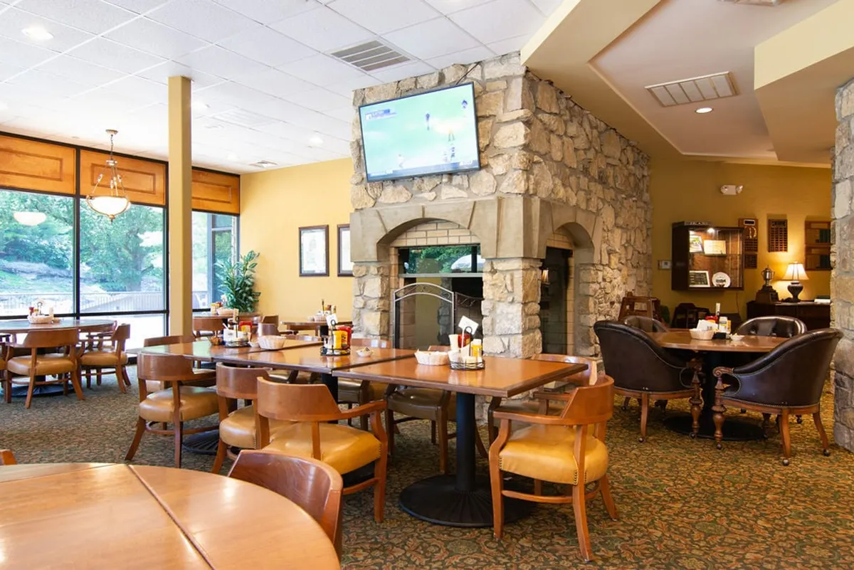 An inviting interior scene of a clubhouse dining area featuring a stone fireplace, large windows offering natural light, several dining tables, and a television—suggesting a relaxed atmosphere for post-golf socializing or meals.